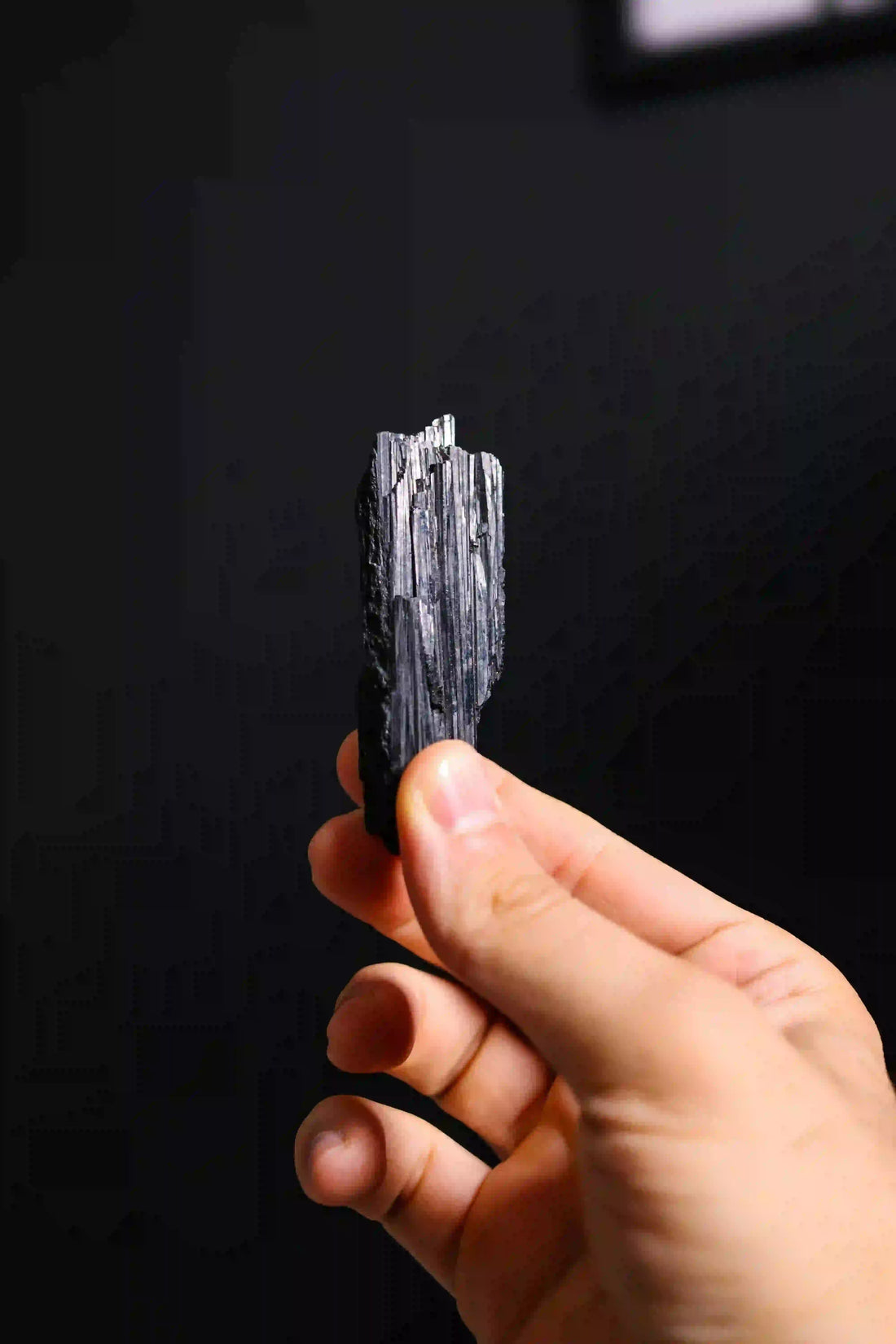 Main tenant un cristal brut de tourmaline noire sur fond sombre, symbole de protection énergétique, d’ancrage et de purification – Aura Cristal